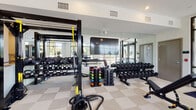 Located at 25 Executive Dr in Stoneham, Massachusetts, this well-appointed fitness center offers a expansive, single-floor layout designed to accommodate a full spectrum of workout needs. The space is bathed in natural light, courtesy of floor-to-ceiling windows that line the perimeter and provide open views of the surrounding greenery, creating an energizing atmosphere for exercise.

The cardio zone features a robust selection of treadmills, elliptical machines, stationary bikes, and rowing machines, all positioned to take advantage of the abundant natural light and outdoor views. Large ceiling fans overhead enhance airflow and comfort throughout the space.

The strength training area is equally well-equipped, with a comprehensive free weights section including a full rack of dumbbells, kettlebells of various sizes, and colorful medicine balls. A multi-functional cable and functional training rig supports TRX suspension training and battle rope workouts. A wide array of selectorized strength machines rounds out the offering for targeted muscle training.

Full-length mirrors along one wall expand the sense of space and allow users to monitor their form. A large-screen television is mounted prominently for added entertainment. The neutral-toned tile flooring, recessed lighting, and clean white walls contribute to the facility's modern, professional aesthetic, making this fitness center a comprehensive and inviting destination for all fitness levels.