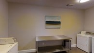 This single-story space presents a clean, understated aesthetic defined by smooth, neutral-toned walls and crisp white baseboards that run along the perimeter. A stainless steel table or work surface commands attention at the center of the frame, suggesting a functional, utilitarian purpose — potentially a commercial, workshop, or utility-oriented environment. The surface's metallic finish reflects light subtly, contributing to the room's practical character. A lower shelf on the steel unit provides additional storage or workspace. Overhead, a ceiling vent indicates a climate-controlled environment. Adding a touch of visual interest to the otherwise minimal setting, a framed canvas featuring a serene, abstract coastal landscape hangs on the wall above the table. The muted tones of teal, grey, and sandy ochre in the artwork contrast softly against the warm beige walls. The overall ambiance is functional yet thoughtfully curated, balancing utilitarian design with a small but deliberate decorative element that brings a sense of calm to the space.