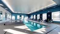This single-level indoor aquatic facility offers a well-appointed and inviting environment designed for both leisure and fitness. The centerpiece is a generously sized rectangular lap pool with crystal-clear turquoise water, framed by smooth light-tiled pool decking that extends throughout the space. Adjacent to the main pool is a separate hot tub or hydrotherapy area, equipped with handrails for easy access, making it suitable for relaxation and therapeutic use.

The interior is finished in a bold deep blue palette on the upper walls, complemented by a clean white tray ceiling with recessed and accent lighting that enhances the bright, airy atmosphere. A row of large windows lines the perimeter, allowing abundant natural light to flood the space and offering views of the surrounding landscape, creating an open and refreshing ambiance.

Along one side of the pool, a row of sleek black lounge chairs with cushions provides a comfortable area for relaxation. The space also features a basketball hoop and a volleyball net positioned over the pool, adding recreational versatility. Overall, the facility balances athletic functionality with a polished, resort-style aesthetic.