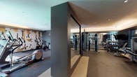 This contemporary fitness center is thoughtfully designed to support a full range of workout routines within a sleek, modern environment. The space is organized into distinct functional zones, allowing for cardio training, strength work, and open-floor exercise without crowding. The cardio area is outfitted with treadmills, elliptical machines, and stationary cycling bikes, all positioned to make use of the natural light streaming in through large floor-to-ceiling glass windows that offer views of the surrounding exterior. A dedicated strength training section houses a multi-functional cable machine system, flanked by floor-to-ceiling mirrors that enhance the sense of space and assist with form. A freestanding dumbbell rack provides a range of free weights for targeted exercises. The open floor area, finished with durable dark rubber flooring, offers room for stretching, balance work, and functional training with stability balls. A built-in shelving unit with warm wood paneling and ambient backlighting serves as a focal design element. The walls are adorned with bold abstract murals in neutral tones with gold accents, contributing an artistic, upscale character to the environment. Recessed and cove lighting throughout creates a well-lit yet refined atmosphere suited for focused training.