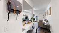 This well-designed single-floor unit offers a smart, efficient layout ideal for student living. The open-concept living and dining area serves as the heart of the space, where a white breakfast bar with bar stools flows seamlessly into a comfortable lounge area. The living room features dark hardwood-style flooring, a flat-screen TV mounted on a sleek media console with a wall-mounted display shelf above, and a cozy area rug that anchors the seating arrangement. Natural light enters through roller-shade windows, while track lighting illuminates the crisp white walls and paneled ceiling overhead.

The unit includes two shared bedrooms, each thoughtfully arranged to accommodate multiple occupants. One bedroom features two twin beds separated by a perforated metal shelving unit that provides both storage and a degree of privacy, along with individual desks and a walk-in closet area with hanging space. The second bedroom similarly offers twin beds, open wardrobe units with hanging rails and drawers, and dedicated study desks — creating a productive personal environment for each resident. Throughout, the design emphasizes clean lines, neutral tones, and practical furnishings, making this a highly functional living space suited to the demands of student life.