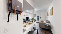 This well-designed single-floor unit offers a smart, efficient layout ideal for student living. The open-concept living and dining area serves as the heart of the space, where a white breakfast bar with bar stools flows seamlessly into a comfortable lounge area. The living room features dark hardwood-style flooring, a flat-screen TV mounted on a sleek media console with a wall-mounted display shelf above, and a cozy area rug that anchors the seating arrangement. Natural light enters through roller-shade windows, while track lighting illuminates the crisp white walls and paneled ceiling overhead.

The unit includes two shared bedrooms, each thoughtfully arranged to accommodate multiple occupants. One bedroom features two twin beds separated by a perforated metal shelving unit that provides both storage and a degree of privacy, along with individual desks and a walk-in closet area with hanging space. The second bedroom similarly offers twin beds, open wardrobe units with hanging rails and drawers, and dedicated study desks — creating a productive personal environment for each resident. Throughout, the design emphasizes clean lines, neutral tones, and practical furnishings, making this a highly functional living space suited to the demands of student life.