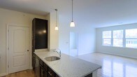 This single-floor residence offers a thoughtfully designed open-concept layout that seamlessly connects the kitchen and living areas. The kitchen is a standout feature, showcasing a generous granite-topped island with a stainless steel undermount sink, dark espresso cabinetry with brushed metal hardware, and a stainless steel dishwasher. Two pendant lights hang above the island, providing warm, focused illumination that complements the kitchen's modern aesthetic. Tall upper cabinetry maximizes vertical storage space, adding to the kitchen's functional appeal. The open layout flows naturally into a bright and spacious living area, where light hardwood flooring extends throughout, adding warmth and continuity to the space. A window in the living area invites natural light, creating an airy and welcoming ambiance. A doorway from the main living space leads to an additional room, suggesting flexible use for a bedroom or home office. The clean white walls and neutral palette throughout provide a versatile backdrop, making the space easy to personalize. Overall, the residence combines practical design with contemporary finishes to deliver a comfortable and stylish single-level living experience.
