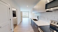 Located in West Lafayette, IN, this thoughtfully designed single-floor unit offers a clean, modern aesthetic throughout. Light-toned hardwood flooring runs continuously across the open-plan layout, lending warmth and visual cohesion to the space.

The kitchen is a standout feature, outfitted with two-tone cabinetry — combining upper light gray units with lower dark wood-finish drawers — and topped with crisp white quartz countertops. A classic white subway tile backsplash, a matte black undermount sink with a matching faucet, and a full suite of stainless steel appliances including a French door refrigerator, electric range with over-the-range microwave, and dishwasher complete this well-appointed space.

The kitchen flows seamlessly into the living area, which benefits from oversized windows on two walls that flood the room with natural light and offer open views of the surrounding West Lafayette streetscape and greenery. The clean white walls and minimalist design create a bright, airy atmosphere throughout.

The bathroom is finished with large-format neutral tile flooring, a white vanity with matte black hardware, and a dark tile-accented shower area, providing a polished, contemporary feel. The unit's efficient single-floor layout, modern finishes, and abundant natural light make it a highly functional and attractive space.