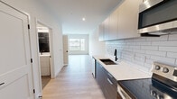 Located in West Lafayette, IN, this thoughtfully designed single-floor unit offers a clean, modern aesthetic throughout. Light-toned hardwood flooring runs continuously across the open-plan layout, lending warmth and visual cohesion to the space.

The kitchen is a standout feature, outfitted with two-tone cabinetry — combining upper light gray units with lower dark wood-finish drawers — and topped with crisp white quartz countertops. A classic white subway tile backsplash, a matte black undermount sink with a matching faucet, and a full suite of stainless steel appliances including a French door refrigerator, electric range with over-the-range microwave, and dishwasher complete this well-appointed space.

The kitchen flows seamlessly into the living area, which benefits from oversized windows on two walls that flood the room with natural light and offer open views of the surrounding West Lafayette streetscape and greenery. The clean white walls and minimalist design create a bright, airy atmosphere throughout.

The bathroom is finished with large-format neutral tile flooring, a white vanity with matte black hardware, and a dark tile-accented shower area, providing a polished, contemporary feel. The unit's efficient single-floor layout, modern finishes, and abundant natural light make it a highly functional and attractive space.