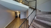 This two-story, 1-bedroom, 1-bathroom unit is thoughtfully designed to maximize its square footage across two distinct levels. The main floor features an open living area finished with warm hardwood-style flooring, neutral gray walls, and clean white baseboards that create a bright, cohesive aesthetic. A compact kitchen is visible from the living space, equipped with dark cabinetry and a black refrigerator, providing functional storage and prep space. Ceiling-mounted lighting illuminates the lower level, while a wall vent ensures comfortable climate control throughout. A carpeted staircase with sleek black metal railings connects the main floor to the upper loft level, adding a striking architectural element to the interior. The upper level features a ceiling fan, suggesting its use as the bedroom or sleeping area, and the open railing design allows sightlines between floors, enhancing the sense of openness and vertical space. The two-level layout efficiently separates living and sleeping areas, making this unit a practical and stylish option for those seeking a loft-inspired living experience.
