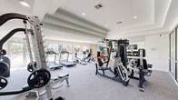 Located at 6336 S 82nd E Ave in Tulsa, Oklahoma, this fitness center offers a comprehensive workout environment within a generously sized, single-floor layout. The space is designed with a clean, modern aesthetic, featuring white walls, recessed lighting, and tray ceiling details that contribute to a bright and open atmosphere. Large floor-to-ceiling windows along one wall flood the interior with natural light while providing direct views of an outdoor pool area, enhancing the overall ambiance.

The main workout floor is finished with neutral gray carpet and is home to a robust selection of cardio equipment, including multiple treadmills, elliptical trainers, stationary bikes, and rowing machines, all arranged to maximize usable floor space. A multi-station cable and strength machine anchors the center of the room, complemented by a dedicated free weight area featuring a full rack of dumbbells, kettlebells, and an adjustable bench positioned beside a wall-length mirror.

A designated functional training zone features a hardwood floor section stocked with medicine balls, stability balls, and additional kettlebells. A wall-mounted television is also present, adding convenience for users during workouts. Glass partition walls on one side offer a visual connection to an adjacent lounge or common area, reinforcing the facility's open and integrated design.