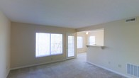 This single-floor space features an open, airy layout designed for comfortable everyday living. The main living area is finished with neutral-toned carpet and painted in a soft beige palette, creating a warm and inviting atmosphere throughout. A large multi-pane window along one wall floods the room with abundant natural light, offering views of surrounding trees and greenery. A glass-panel French door provides direct access to the outdoors, further enhancing the connection to the natural environment and allowing additional light to pour in.

Adjacent to the living area, a pass-through counter connects to a separate kitchen or dining space, making the layout practical for entertaining or daily use. This adjoining area is equipped with a ceiling fan, contributing to airflow and comfort. The pass-through design maintains an open feel between the spaces while still offering a degree of visual separation. White trim and baseboards throughout provide clean, crisp contrast against the neutral walls and carpeted floors. Overall, the space presents a functional and well-lit environment with a straightforward, comfortable design suitable for a range of uses.