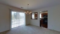 This single-floor residence offers a practical and comfortable layout designed for everyday living. The dining area serves as a central gathering space, highlighted by an elegant multi-candle chandelier that draws the eye upward and adds a touch of classic charm. Large sliding glass doors with vertical blinds flood the room with abundant natural light and provide direct access to an outdoor patio area, offering pleasant views of the surrounding greenery. The neutral-toned carpet and light-colored walls create a clean, versatile backdrop throughout the living and dining space. A pass-through window with a dark wood frame connects the dining area to the kitchen, enhancing the open feel between the two spaces. The kitchen is well-appointed with rich, warm-toned wood cabinetry, black appliances including a range and built-in microwave, and tiled flooring that contrasts neatly with the carpeted dining area. The overall layout promotes a seamless flow between cooking and dining spaces, making it well-suited for both daily use and casual entertaining. The home's straightforward, single-level design ensures easy accessibility and a cohesive living experience throughout.