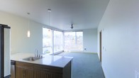 This contemporary single-floor space is defined by its clean lines, neutral palette, and an abundance of natural light. The open-plan layout seamlessly connects the kitchen and living area, creating a cohesive and airy environment. The kitchen features a sleek island with a light-toned countertop, an undermount sink, and dark wood cabinetry, providing ample workspace and storage. A pendant light fixture above the island adds a refined focal point. The living area is finished with low-pile carpeting and anchored by a ceiling fan, ensuring year-round comfort. The most striking feature of the space is the expansive corner window arrangement, which floods the interior with natural light and frames panoramic views of the surrounding urban landscape and tree canopy beyond. The smooth white ceilings and clean wall surfaces maintain a minimalist aesthetic throughout, allowing the views and natural light to take center stage. A wall-mounted heater beneath the windows provides efficient climate control. The overall ambiance is bright, open, and modern, making this a highly functional and visually appealing space suited to a variety of uses.