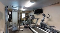 This fitness room offers a practical and well-equipped space for residents or guests to maintain their workout routines. The room features a selection of cardio equipment, including at least two treadmills and an elliptical machine, catering to a range of cardiovascular exercise preferences. A multi-function cable weight station is positioned along one wall, providing options for strength and resistance training. A wall-mounted flat-screen television adds entertainment value during workouts.

The room's mirrored wall visually expands the space while serving the functional purpose of allowing users to monitor their form during exercise. Overhead fluorescent lighting ensures the space is well-lit throughout. A textured ceiling and neutral-toned walls contribute to the room's utilitarian aesthetic. A small portable fan is also present, offering additional air circulation during use. A window allows a modest amount of natural light to enter the space, complementing the artificial lighting. The overall layout is efficient, maximizing the use of available floor space while keeping the environment focused and functional for fitness purposes.