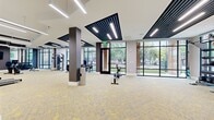 This contemporary fitness center offers a well-appointed, single-floor layout designed to accommodate a full range of workout needs. The space is divided into distinct functional zones, including a cardio area outfitted with treadmills, ellipticals, and stationary bikes, as well as a strength training zone featuring cable machines, free weights, dumbbells, medicine balls, and benches. A generous open floor area provides ample room for stretching and functional training exercises.

Architecturally, the facility is defined by its striking ceiling design, which incorporates bold geometric recessed panels with dark slatted accents and integrated linear LED lighting, creating a sleek, modern aesthetic. Expansive floor-to-ceiling windows and glass-paneled walls line multiple sides of the space, flooding the interior with abundant natural light and offering views of the surrounding outdoor greenery and streetscape.

The flooring throughout features stylish patterned carpet tiles in gray and yellow tones, adding a vibrant yet professional touch. Large wall-mounted mirrors enhance the sense of openness in the cardio section, while dark structural columns punctuate the space with an industrial-modern contrast. Additional amenities include a water fountain station and restroom access, ensuring convenience for all users. The overall ambiance is bright, energetic, and polished.