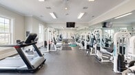 Located at 200 Atrium Way in Columbia, South Carolina, this fitness center offers a dedicated, single-room exercise space designed to support a comprehensive workout experience. The room is thoughtfully laid out with durable rubber flooring, providing a safe and functional surface for a wide range of physical activities.

The space is equipped with an impressive array of fitness machines, including treadmills, a stationary bike, a cable/weight machine, and multiple strength-training stations, accommodating both cardio and resistance training. Floor-to-ceiling mirrors line one wall, visually expanding the room while allowing users to monitor their form during workouts.

Overhead, recessed fluorescent panel lighting and a ceiling fan ensure the space remains well-lit and properly ventilated. Multiple multi-pane windows along the walls invite ample natural light and offer views of the surrounding outdoor greenery, contributing to an open and energizing atmosphere. Crown molding and white trim details along the walls and ceilings lend a polished, refined aesthetic to the room.

Entry is provided through elegant French doors, adding an architectural touch that complements the clean, neutral color palette of soft gray and white tones throughout. A wall-mounted flat-screen television adds a modern amenity for entertainment during workouts. The overall ambiance is bright, clean, and well-maintained, making it an inviting environment for fitness enthusiasts.