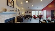 This single-story clubhouse is thoughtfully designed to serve both community gathering and administrative functions. The main lounge area is the centerpiece of the space, boasting vaulted ceilings with recessed lighting and a ceiling fan that contribute to an open, airy atmosphere. A tile-surround fireplace anchors one wall, complemented by a built-in shelving unit displaying decorative accents. Comfortable dark upholstered sofas and accent chairs in warm red tones are arranged around a glass-top coffee table, creating an inviting seating area. A bold red accent wall with a wall-mounted television adds a modern focal point, while large windows with blinds allow natural light to filter throughout.

Adjacent to the lounge is a dedicated leasing or administrative office area, featuring a rich mahogany executive desk, a computer workstation, and direct access through a private door. The space is finished with grey carpeting and neutral grey walls, maintaining a professional yet welcoming atmosphere.

A separate business center provides residents with shared computing resources, including two desktop computer stations and a multifunction printer, all set on a clean, light-toned workbench—ideal for productivity needs.