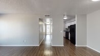This single-story unit offers a practical and straightforward layout connecting a living area, kitchen, bedrooms, and bathroom. The living room serves as the main gathering space, featuring dark hardwood-style flooring that extends throughout the common areas, neutral gray walls, and a textured ceiling. A window along one wall allows natural light to brighten the room, while a front entry door provides a clear sense of arrival.

The kitchen is positioned adjacent to the living area and is equipped with white cabinetry offering generous upper and lower storage, blue-toned laminate countertops, a stainless steel sink, and black appliances including a refrigerator, range, and dishwasher. A hallway connects the main living spaces to the private areas of the unit.

The bedrooms are carpeted in neutral tones and feature textured ceilings and white baseboards. Each bedroom includes closet space with bifold doors, providing practical storage. The bathroom is accessible from the hallway and includes a tub-shower combination with white tile surround, a toilet, and a vanity with a countertop, all set on hardwood-style flooring. The overall layout is efficient, with each room clearly defined and easy to navigate.