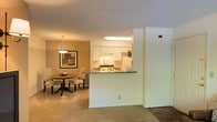 This single-floor space is thoughtfully designed with an open-concept layout that seamlessly connects the kitchen, dining area, and living space. The kitchen is positioned behind a half-wall peninsula, which serves as a natural divider while maintaining visual openness throughout. White cabinetry lines the kitchen walls, complemented by a light countertop surface that extends to the peninsula overhang. Overhead, a skylight-style fixture brightens the kitchen, ensuring the space remains well-lit. The dining area is carpeted and features warm, golden-toned walls that create an inviting ambiance. A pendant light hangs above a round wooden dining table surrounded by upholstered chairs, adding a classic and refined touch. A framed piece of wall art anchors the dining nook, enhancing the warmth of the space. The flooring transitions from carpet in the living and dining areas to a harder surface near the entryway, adding visual contrast. The textured ceiling and neutral tones throughout contribute to a cohesive, comfortable atmosphere well-suited for everyday living.