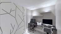 This single-floor space showcases a contemporary, minimalist aesthetic that is both functional and visually refined. The workspace is defined by a clean, neutral palette of soft grays and whites, creating a calm and focused environment well-suited for productivity.

A built-in desk spans the width of the room, providing generous workspace accommodating dual monitor setups alongside peripheral equipment such as a printer. Above the desk, a floating wall-mounted shelf offers additional storage or display space while maintaining the room's streamlined appearance. The recessed lighting integrated into the ceiling contributes to a clean, uncluttered look while providing consistent, even illumination throughout the space.

A standout design feature is the large decorative wall panel on the left side of the room, adorned with an abstract branching pattern rendered in gray against a light background. This artistic element introduces visual interest and texture without disrupting the overall minimalist character of the space.

Ergonomic mesh-back task chairs complement the modern workstations, reinforcing the office's practical yet design-conscious approach. The overall ambiance is professional, orderly, and thoughtfully composed.