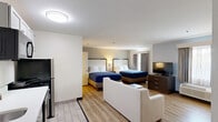 Located at 835 Upper Union St in Franklin, MA, this compact yet efficiently designed suite offers a thoughtfully organized open-plan layout across a single floor. The main living area seamlessly integrates a fully equipped kitchenette featuring white shaker-style cabinetry, a stainless steel refrigerator, microwave, stovetop, dishwasher, and a sink with ample counter space — ideal for extended stays or self-sufficient living. Light wood-look flooring flows throughout the space, complementing the clean, neutral color palette of white and soft grey tones. The living area is furnished with a sofa and coffee table, providing a comfortable lounge space, while the sleeping area accommodates two beds dressed in crisp blue and white linens. A dresser with a flat-screen TV, a dedicated desk with seating, and a wall-unit climate control system round out the room's functionality. A large closet with shelving and hanging space adds practical storage. Natural light filters in through windows dressed with layered curtains. The private bathroom is finished with light tile flooring, a glass-enclosed shower, a floating vanity with a white countertop, and clean white walls accented by modern fixtures.