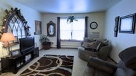 This single-floor space offers a warm and comfortable living environment with a thoughtfully arranged interior. The living area serves as the centerpiece of the space, featuring wall-to-wall carpeting that adds a soft, cozy feel underfoot. A large window dressed with dark curtains allows natural light to flood the room, creating a bright and welcoming atmosphere. A bold patterned area rug in brown and teal tones anchors the seating area, adding a striking visual element to the room. The living space accommodates a curved sectional sofa adorned with decorative pillows, providing ample seating and a relaxed, casual ambiance. A wall-mounted clock and an ornate framed mirror add character to the walls, while framed artwork contributes to the room's personalized charm. Greenery in the form of potted plants, including a hanging plant near the window, brings a touch of nature indoors and enhances the overall freshness of the space. A television setup on a media console rounds out the living area, making it well-suited for both relaxation and entertainment. The baseboard heating system along the walls ensures comfort throughout the seasons.