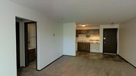 This single-floor space features an open-concept layout that connects the living area to the kitchen, creating a functional and cohesive floor plan. The living area is finished with wall-to-wall carpet, providing a comfortable and quiet underfoot feel, while dark baseboards offer a subtle contrast against the white walls throughout.

The kitchen is positioned at the rear of the open space and is outfitted with dark wood cabinetry spanning both upper and lower storage, offering ample cabinet space. White appliances, including a range and what appears to be a refrigerator, are integrated neatly beneath the cabinetry. A flush-mounted ceiling light illuminates the kitchen area, and an under-cabinet range hood is installed above the stovetop.

To the left of the main living area, a partially visible alcove features additional cabinetry and what appears to be a closet or utility space with a hanging rod. The flooring in this transitional zone shifts to a hard surface tile, distinguishing it from the carpeted living space. The textured ceiling is consistent throughout, and a dark entry door is visible near the kitchen, indicating a direct exterior access point.