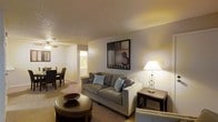 This single-floor space is designed with an open, functional layout that connects the living and dining areas seamlessly. The living room serves as the central gathering space, anchored by a plush sofa adorned with decorative pillows in neutral tones. A round wooden coffee table adds warmth to the seating area, while a matching side table with a classic lamp provides ambient lighting. A bold piece of framed wall art adds a focal point above the sofa. The textured ceiling and warm, neutral wall tones create a cozy atmosphere throughout. Soft carpeted flooring extends across the living and dining areas, contributing to the overall comfort of the space. The dining area, visible from the living room, features a modest table with upholstered chairs. A ceiling fan overhead enhances air circulation. A hallway visible in the background leads to additional rooms, including what appears to be a well-lit bathroom. The space benefits from a warm, inviting ambiance created by layered lighting, including both overhead and accent sources, making it ideal for relaxed everyday living.