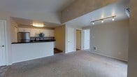 This single-floor space offers an open-concept layout that seamlessly connects the kitchen and living areas, creating a functional and inviting environment. The kitchen is equipped with stainless steel appliances, including a range and an over-the-range microwave, complemented by white cabinetry and dark countertops. A peninsula-style counter provides a natural divider between the kitchen and the main living area, adding both workspace and a casual dining option. The flooring transitions from hardwood in the kitchen to plush, neutral-toned carpet throughout the spacious living area, contributing to a warm and comfortable atmosphere. Vaulted ceilings rise prominently over the living space, enhancing the sense of openness and volume. Track lighting along the ceiling provides focused illumination, while the overall neutral color palette of beige and white tones keeps the space feeling bright and clean. Two interior doors visible from the main living area lead to additional rooms, suggesting a practical and well-organized floor plan. The space combines modern finishes with a straightforward, accessible design suited for everyday living.