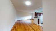 Located at Wildwood Gardens in Port Washington North, NY, this single-floor residence offers a straightforward and functional layout with a clean, understated aesthetic throughout. Warm-toned hardwood flooring extends across the living areas and bedrooms, lending a cohesive and inviting feel to the interior.

The living area is open and bright, with neutral white walls and baseboard heating running along the perimeter. The space connects naturally to the kitchen, which is separated by a partial wall and features rich, dark wood cabinetry, dark granite-style countertops, tile flooring, a gas range, refrigerator, and a window that brings in natural light. The galley-style configuration makes efficient use of the available space while providing ample cabinet storage.

The bedroom accommodates a comfortable living area with a window fitted with an air conditioning unit, hardwood floors, and baseboard heating. Closet space is available, with built-in shelving providing additional storage options.

The bathroom is finished with light beige tile, a decorative mosaic accent border, and a full bathtub enclosed by sliding glass shower doors, with a small window for ventilation. The overall space presents a clean and practical environment well-suited for everyday living.