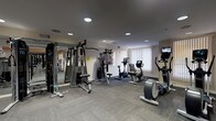 This fitness center offers a thorough and well-organized workout environment spread across a single floor. The space is divided into distinct zones, each designed to accommodate different training needs. The strength training area features a robust selection of Precor selectorized machines, including chest press, shoulder press, lat pulldown, and cable systems, all arranged to allow comfortable movement between stations. Full-length mirrors line portions of the walls, enhancing the sense of openness and allowing users to monitor their form. A dedicated functional fitness zone houses an impressive array of accessories, including stability balls, BOSU trainers, resistance bands, kettlebells, and a comprehensive rack of dumbbells. The cardio section is equipped with treadmills, elliptical trainers, stationary bikes, and a rowing machine, several of which feature individual screens for entertainment. The flooring throughout consists of durable, cushioned carpet tiles suited for high-traffic fitness use. Recessed lighting illuminates the space evenly, while large windows allow natural light to filter in. A water cooler station is conveniently positioned near the entrance. The overall ambiance is clean, professional, and well-maintained, making it a fully functional facility for a broad range of fitness routines.