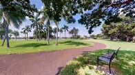 This inviting outdoor space captures the essence of a well-maintained tropical park, offering a serene and scenic environment ideal for leisure and recreation. A gently curving pathway, finished in a rich terracotta-toned rubberized surface, winds through the landscape, guiding visitors past a canopy of mature trees and clusters of towering royal palms that define the character of the space. The lush, well-kept lawn stretches expansively between the pathway and the waterfront, where a graceful pedestrian bridge can be spotted in the distance, adding a charming architectural accent to the natural setting. Seating is thoughtfully integrated into the environment, with a classic park bench positioned along the path, inviting visitors to pause and take in the surroundings. The open sky above allows abundant natural light to filter through the tree canopy, casting dappled shade across the grounds. The overall ambiance is calm and refreshing, with the combination of tropical flora, open green space, and nearby water creating a picturesque and tranquil retreat. The landscape reflects the vibrant, sun-drenched character typical of South Florida's outdoor spaces, making it a welcoming destination for walking, relaxing, and enjoying the natural environment.