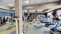 Located at 4209 Lassiter Mill Rd in Raleigh, NC, this well-appointed fitness center offers a comprehensive workout environment across a single, open floor. The space is designed with a modern aesthetic, featuring a bold two-tone color scheme of deep navy blue and warm taupe on the walls, complemented by a distinctive multi-colored geometric carpet in shades of teal, beige, and gray. A drop-panel ceiling with recessed lighting and ceiling fans ensures the space remains bright and comfortable throughout.

The cardio zone is positioned along a windowed wall that floods the area with natural light and provides views of an outdoor pool and landscaping, creating an energizing atmosphere. This section is equipped with a row of True-brand treadmills, elliptical trainers, and recumbent bikes, each outfitted with personal screens. The strength training area features a robust selection of selectorized cable machines, a multi-functional cable crossover unit, and a dedicated free weights section with a full dumbbell rack and medicine ball storage. Additional functional training tools, including stability balls, resistance equipment, and a SmartMat, are neatly organized along the walls. Large mirrors throughout the space enhance the sense of openness and support proper form during workouts.