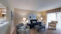This single-floor residence offers two bedrooms and one bathroom, designed with a practical and comfortable layout. The living room serves as the central gathering space, finished with wall-to-wall carpeting and neutral beige walls that create a warm, inviting atmosphere. A window along one wall allows natural light to filter into the room, brightening the space throughout the day. The room comfortably accommodates a full seating arrangement, as demonstrated by the navy sofa, upholstered armchair, and ottoman centerpiece visible in the space. Decorative framed botanical artwork adorns the walls, contributing to the cohesive, calming aesthetic. Soft lighting from table lamps supplements the natural light, ensuring a well-lit environment at all times. The clean, simple lines of the architecture keep the focus on functionality and livability. With two bedrooms, the residence provides flexibility for guests, a home office, or additional living needs. The single-bathroom layout is complemented by the efficient, single-story floor plan, making the space easily navigable and accessible throughout.