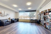 The Social Room at 1032 King St W in Kingston, Ontario, is a thoughtfully designed communal gathering space that combines comfort and functionality in a single open-concept floor. The living area is anchored by warm hardwood-style flooring and features a seating arrangement of leather armchairs paired with a side table, creating an inviting lounge atmosphere. Decorative multi-panel artwork adorns the neutral-toned walls, adding a touch of warmth to the space. A standout feature is the expansive floor-to-ceiling window along one wall, fitted with roller blinds, which floods the room with natural light and offers pleasant views of the surrounding outdoor landscape. Adjacent to the seating area, a well-stocked bookshelf library lines an entire wall, providing a curated collection of books and games for residents to enjoy. The kitchen area is clean and fully equipped, featuring white shaker-style cabinetry extending to the ceiling, stainless steel appliances including a range and refrigerator, a dishwasher, a microwave, and a double sink — all finished with light tile flooring. Flush-mount ceiling lighting throughout ensures the space remains bright and functional at all hours, making this Social Room an ideal hub for community gatherings, reading, and casual entertaining.