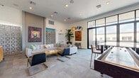 Located at 4333 N 6th Dr in Phoenix, Arizona, this single-floor community lounge known as "Curve" offers a generously sized, open-plan gathering space designed for comfort and social interaction. The interior is defined by its modern aesthetic, featuring plush sectional sofas, mixed seating arrangements, and an eclectic collection of bold, colorful artwork displayed throughout — including large-scale paintings and a distinctive decorative lattice screen partition that adds architectural depth.

The main lounge area benefits from soaring ceilings and abundant natural light, drawn in through an expansive floor-to-ceiling glass wall and sliding door system that opens directly onto an outdoor balcony. The balcony is furnished with high-top dining sets and lounge seating, offering open views of the Phoenix skyline under wide desert skies.

Inside, a dedicated media corner features wall-mounted flat-screen televisions above a mid-century-style wood credenza, perfect for casual viewing. A dining and work area nearby is lit by a statement petal-shaped pendant light, accompanied by varied seating and a wall-mounted display screen. A teal accent wall anchors the far end of the space, adding a pop of color alongside pendant lighting. Tall indoor plants, recessed lighting, and plush carpeting complete the warm, inviting atmosphere throughout.