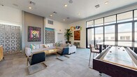 Located at 4333 N 6th Dr in Phoenix, Arizona, this single-floor community lounge known as "Curve" offers a generously sized, open-plan gathering space designed for comfort and social interaction. The interior is defined by its modern aesthetic, featuring plush sectional sofas, mixed seating arrangements, and an eclectic collection of bold, colorful artwork displayed throughout — including large-scale paintings and a distinctive decorative lattice screen partition that adds architectural depth.

The main lounge area benefits from soaring ceilings and abundant natural light, drawn in through an expansive floor-to-ceiling glass wall and sliding door system that opens directly onto an outdoor balcony. The balcony is furnished with high-top dining sets and lounge seating, offering open views of the Phoenix skyline under wide desert skies.

Inside, a dedicated media corner features wall-mounted flat-screen televisions above a mid-century-style wood credenza, perfect for casual viewing. A dining and work area nearby is lit by a statement petal-shaped pendant light, accompanied by varied seating and a wall-mounted display screen. A teal accent wall anchors the far end of the space, adding a pop of color alongside pendant lighting. Tall indoor plants, recessed lighting, and plush carpeting complete the warm, inviting atmosphere throughout.