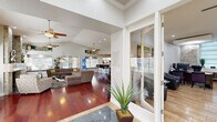 This well-appointed single-story space offers a thoughtfully designed open floor plan that blends comfort and sophistication throughout. The entry foyer makes a striking first impression with travertine tile flooring, a stacked-stone accent wall, and a bold piece of integrated artwork framed in mirrored panels. Adjacent to the entry is a professional reception area featuring light tile flooring, recessed lighting, a dedicated workspace, and a comfortable seating arrangement — ideal for welcoming guests or clients.

The expansive main living area is defined by soaring vaulted ceilings, gleaming rich hardwood floors, and abundant natural light filtering through multiple windows with custom treatments. A central stacked-stone fireplace with a wood mantle serves as the focal point of the room, flanked by built-in display shelves and framed by two ceiling fans. The generous layout accommodates large sectional seating and provides an open sightline throughout the space.

The kitchen is equipped with warm wood cabinetry, granite countertops, a curved breakfast bar with modern bar seating, black appliances, and a double basin sink beneath a window overlooking the exterior. A built-in window bench with cushioned seating adds a charming and functional nook. The cohesive design palette of neutral tones, stone textures, and warm wood finishes creates an inviting and polished ambiance throughout.