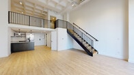 This two-floor loft offers a striking blend of industrial character and modern design. The main level welcomes you with a generous open-plan living area boasting soaring ceilings with exposed structural beams and ductwork painted white, lending an airy, urban feel. Light wood flooring runs throughout, complementing the bright, white walls. A sleek kitchen anchors the space, featuring a two-tone cabinet scheme with dark lower cabinets and crisp white upper cabinets, a classic white subway tile backsplash, stainless steel appliances, and a peninsula with an integrated sink and matte black faucet, illuminated by pendant lighting overhead. A bold black metal staircase with matching railings leads to the upper level, where an open mezzanine overlooks the living area below. Large floor-to-ceiling windows on the upper floor flood the space with natural light and frame urban rooftop views. A private outdoor terrace, accessible via glass sliding doors, provides a spacious retreat with city views. The bathroom is finished in clean white tile with a soaking tub and matte black fixtures and hardware. A dedicated laundry area houses a stacked LG washer and dryer unit, adding everyday convenience to this thoughtfully designed urban loft.