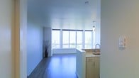 This single-floor space features a bright, open-concept layout that seamlessly integrates the living area with the kitchen. The most striking element is the expansive bank of floor-to-ceiling windows along the far wall, which flood the interior with natural light and offer panoramic views of the surrounding cityscape and greenery. Roller blinds are installed to provide adjustable light control throughout the day.

The living area is finished with smooth hardwood-style flooring in a cool grey-brown tone, lending the space a sleek, contemporary feel. White walls and a clean white ceiling contribute to an airy, minimalist ambiance throughout.

The kitchen is positioned along one side of the open layout and is defined by warm wood cabinetry with simple, modern hardware. The countertops feature a light stone or stone-look surface, and a stainless steel sink with a curved gooseneck faucet serves as a functional centerpiece. A pendant light fixture hangs nearby, adding a subtle decorative accent. The kitchen flows naturally into the main living area, making the space feel cohesive and well-suited for everyday living and entertaining.