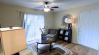 This single-story living space is designed with both comfort and functionality in mind. The living area serves as the focal point of the home, featuring warm hardwood-style flooring that adds a sense of depth and cohesion throughout. A large window dressed with vertical blinds allows abundant natural light to filter into the room, creating a bright and airy ambiance while offering a view of the outdoors. A ceiling fan with an integrated light fixture is centrally positioned, providing both practical air circulation and supplemental lighting. The space is laid out to accommodate a comfortable seating arrangement, with room for a round coffee table and accent furniture, including a compact shelving unit. A textured ceiling adds a subtle architectural detail overhead. The neutral wall tones create a versatile backdrop that complements a variety of décor styles. Overall, the living area strikes a balance between openness and coziness, making it a practical and inviting space for everyday living.