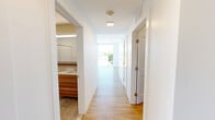 Located at 2–10 Mackin St in Boston's Brighton neighborhood, this contemporary 2-bedroom, 2-bathroom residence offers a thoughtfully designed single-floor layout with clean, modern finishes throughout. Light wood-tone flooring runs continuously across the living areas and bedrooms, complementing the bright white walls that give the space an open, airy feel.

The kitchen is a standout feature, showcasing a large quartz waterfall island with an undermount sink, stainless steel appliances including a refrigerator, range, and microwave, warm walnut-toned upper cabinetry, and open floating wooden shelves. A tall floor-to-ceiling window floods the kitchen with natural light and provides views of the surrounding Boston neighborhood.

The primary bedroom is generously sized and benefits from expansive floor-to-ceiling windows that bring in abundant sunlight and offer elevated city views. The second bedroom is also well-proportioned with its own closet access and wood flooring. Both rooms are finished with crisp white walls and clean lines.

The two bathrooms feature neutral stone-look tile flooring. One includes a walk-in shower with a modern vanity featuring warm wood cabinetry, while the second offers a full tub-shower combo. A dedicated laundry room houses a stacked washer and dryer unit, adding everyday convenience to this efficiently designed residence.