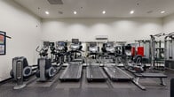 This fitness center in New Orleans, Louisiana, offers a thoroughly equipped workout environment spread across a single floor. The space is organized into distinct functional zones, providing a structured experience for both cardio and strength training. The cardio area features multiple treadmills, elliptical trainers, and stationary bikes, each equipped with individual monitor screens for entertainment during workouts. The flooring throughout is a durable rubber gym surface, providing stability and impact absorption. Full-length mirrors line the walls, visually expanding the space while allowing users to monitor their form.

The strength training zone includes a variety of cable and resistance machines, along with a dedicated free weights area housing an extensive dumbbell rack with weights ranging across multiple increments, accompanied by adjustable benches. Additional cable machines and multi-functional weight stations provide options for a wide range of exercises. Instructional fitness charts are displayed on the walls throughout the facility. Recessed lighting illuminates the space evenly, and a wall-mounted air conditioning unit supports climate control. The overall environment is clean, organized, and designed to support a comprehensive fitness routine.