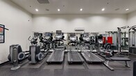 This fitness center in New Orleans, Louisiana, offers a thoroughly equipped workout environment spread across a single floor. The space is organized into distinct functional zones, providing a structured experience for both cardio and strength training. The cardio area features multiple treadmills, elliptical trainers, and stationary bikes, each equipped with individual monitor screens for entertainment during workouts. The flooring throughout is a durable rubber gym surface, providing stability and impact absorption. Full-length mirrors line the walls, visually expanding the space while allowing users to monitor their form.

The strength training zone includes a variety of cable and resistance machines, along with a dedicated free weights area housing an extensive dumbbell rack with weights ranging across multiple increments, accompanied by adjustable benches. Additional cable machines and multi-functional weight stations provide options for a wide range of exercises. Instructional fitness charts are displayed on the walls throughout the facility. Recessed lighting illuminates the space evenly, and a wall-mounted air conditioning unit supports climate control. The overall environment is clean, organized, and designed to support a comprehensive fitness routine.
