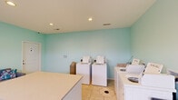 Located at 2759 Towne Blvd in Middletown, Ohio, this communal laundry room is a fully functional, single-floor facility designed to serve residents with convenience and comfort. The room is finished with light aqua/mint-green walls that lend a clean, airy atmosphere, complemented by neutral tile flooring throughout. Recessed ceiling lighting ensures the space is well-lit at all times.

The laundry area is equipped with multiple top-loading washing machines and front-loading dryers, arranged along the walls to maximize floor space. Spacious folding counters provide ample surface area for sorting and folding laundry. A rolling laundry cart is available for ease of transport, and a waste bin is conveniently positioned nearby.

A glass-paneled exterior door allows natural light to filter into the room while offering a view of the surrounding grounds. A horizontal clerestory window on the adjacent wall further enhances the natural light. The entry area features a welcome mat and clearly marked exit signage for safety.

Adding a residential touch, a cushioned sofa with decorative pillows is positioned in a corner of the room, giving residents a comfortable place to wait. Framed wall art adds a subtle decorative element. The overall layout is practical yet inviting, making routine laundry tasks a more pleasant experience.