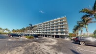 This mid-rise building presents a striking coastal aesthetic, characterized by its crisp white exterior and a uniform arrangement of private balconies extending across each floor. The structure rises several stories, offering residents elevated vantage points and the potential for expansive outdoor living on individual terraces framed by contemporary railings. The ground level is dedicated to covered parking, providing convenient and protected vehicle storage directly beneath the building — a practical and well-integrated architectural feature. The building's clean, modernist lines are complemented by the lush tropical landscaping that surrounds the property, with tall, mature palm trees adding a distinctly South Florida character to the setting. Large windows along the façade suggest bright, light-filled interiors that benefit from the abundant natural sunlight of the region. The overall ambiance is one of relaxed coastal living, with the building's design prioritizing outdoor access and openness at every level. The well-maintained exterior and spacious surface parking lot reflect a community-oriented environment well suited to a warm-weather lifestyle. The surrounding environment, marked by blue skies and verdant palms, reinforces the resort-like atmosphere that defines this property.