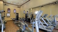 This single-level fitness center is thoughtfully designed to accommodate a comprehensive workout experience. The space features warm beige walls accented by rich crown molding, lending a refined aesthetic uncommon in typical gym environments. Wood-style flooring runs throughout, providing both durability and visual warmth underfoot.

The room is generously proportioned, with elevated ceilings that contribute to an open, airy atmosphere. Ceiling fans are strategically positioned to maintain comfortable airflow during workouts. Recessed lighting supplements the natural light that filters in through arched, shuttered windows — a distinctive architectural detail that adds character to the space.

Floor-to-ceiling mirrors line one wall, visually expanding the room while allowing users to monitor their form during exercise. The cardio section is well-appointed with elliptical trainers and treadmills, while a dedicated strength training area includes a multi-station cable machine and a full dumbbell rack with a wide range of weights. Rubber floor mats are placed beneath the equipment for protection and stability.

Overall, the fitness center combines practical functionality with polished design, creating an inviting environment that encourages consistent and effective training.