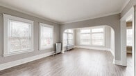 Located at 12933 Clifton Blvd in Lakewood, Ohio, this 2-bedroom, 1-bathroom residence showcases a blend of classic character and functional design across a well-organized single floor. The spacious living room immediately impresses with its crown molding, neutral gray walls, light wood-style flooring, and an elegant arched passageway flanked by large multi-pane windows that flood the room with natural light. Vintage cast-iron radiators are present throughout, adding a charming period detail while providing reliable warmth. The separate dining room offers additional living space with its own natural light and flows conveniently into the kitchen. The kitchen is equipped with warm oak cabinetry, dark countertops, a subway tile backsplash, a double sink, and a full-size refrigerator, with a door providing direct exterior access. Both bedrooms are comfortably sized, featuring crown molding, neutral tones, and their own windows with neighborhood views — one with hardwood-style flooring and one with plush carpet. Two closets offer ample storage. The bathroom includes a full soaking tub with shower and a window for natural ventilation. A connecting hallway ties all rooms together for a practical, cohesive layout.