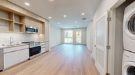 Located along the iconic Sunset Boulevard in Los Angeles, this thoughtfully designed 1-bedroom, 2-bathroom residence offers a clean, contemporary aesthetic throughout. Light-toned hardwood flooring flows seamlessly across the entire space, creating a warm and cohesive atmosphere.

The open-concept kitchen and living area serves as the heart of the home. The kitchen is outfitted with flat-front light wood cabinetry, open shelving, white tile backsplash, stainless steel appliances including a gas range, dishwasher, over-the-range microwave, and a full-size refrigerator. Recessed lighting illuminates the space efficiently.

The living area benefits from large windows and a glass-panel door that opens to a private balcony, offering lush views of swaying palm trees and the modern building courtyard below — a quintessentially Los Angeles backdrop.

The bedroom is a bright, serene retreat with a large window framing palm tree views and abundant natural light. An adjacent walk-in closet provides generous built-in shelving and hanging space, offering excellent organization potential.

The primary bathroom is finished with large-format grey tile flooring, a dual-sink vanity with wood cabinetry, backlit rounded mirrors, and a glass-enclosed walk-in shower. The second bathroom provides additional convenience. A clean hallway with recessed lighting connects all areas of the residence.