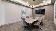 Located at 1703 Rockhill Rd in McKinney, TX, this refined conference room offers a professional and well-appointed setting designed for productive meetings. The room is defined by a distinctive tray ceiling with integrated cove lighting and recessed fixtures, as well as a sleek linear pendant light suspended above the central conference table, contributing to a warm and focused ambiance.

A large wall-mounted flat-screen display is set against a textured herringbone-patterned accent wall, creating a functional focal point ideal for presentations and collaborative sessions. The light wood conference table comfortably accommodates up to eight, surrounded by upholstered ergonomic chairs in neutral tones that complement the room's sophisticated palette.

The walls are finished in crisp white and adorned with framed abstract artwork, adding a curated aesthetic to the space. Patterned low-pile carpeting grounds the room with subtle visual interest. A full-height glass partition with a black-framed door opens the room to the broader facility, allowing visibility and natural light to filter through while maintaining acoustic separation. The overall design balances modern professionalism with thoughtful decorative detail, making it a versatile and inviting space for meetings, collaboration, and presentations.