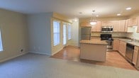 This single-floor residence showcases an open-concept layout that seamlessly connects the kitchen, dining, and living areas, creating a bright and functional environment. The kitchen is well-appointed with stainless steel appliances, including a refrigerator, range, and microwave, all complemented by light wood cabinetry and a granite-topped center island. A pendant light fixture hangs above the island, adding a stylish focal point to the space. The kitchen transitions smoothly into the living area, where warm hardwood flooring gives way to plush carpeting, clearly defining the separate zones while maintaining a cohesive flow. Neutral-toned walls in soft yellow and gray tones lend a welcoming ambiance throughout. Natural light fills the space through multiple windows dressed with vertical blinds, and a glass-paneled door at the far end of the living area provides access to the exterior, enhancing the sense of openness. Recessed and flush-mount ceiling lights ensure the space remains well-lit throughout. The overall design balances practicality with comfort, making it a versatile and inviting single-floor living space suited for everyday living.