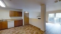This single-floor space offers a functional and straightforward layout designed for comfortable everyday living. The kitchen is outfitted with rich, warm-toned wood cabinetry — featuring both upper and lower cabinet storage — paired with a clean countertop workspace and a dishwasher. Light hardwood-style flooring runs throughout, lending a cohesive and inviting feel to the interior. A pendant light fixture hangs centrally, providing focused illumination over the kitchen and dining transition area. A half-wall partition with a small built-in shelf subtly separates the kitchen from the adjacent living area, maintaining an open and airy connection between the two spaces. The living area, partially visible beyond the divider, benefits from natural light that brightens the room. Neutral white walls throughout keep the space feeling clean and adaptable to a variety of interior styles. A solid wood interior door provides access to an additional room or exterior entry. Overall, the space presents a practical, well-organized floor plan with classic finishes that create a warm and livable atmosphere.