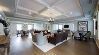Located at 1000 Providence Drive in Islip Terrace, New York, this two-story property showcases a thoughtfully designed interior with notable architectural character. The main gathering area is defined by striking coffered ceilings with crisp white molding, lending a sense of height and grandeur to the space. Recessed lighting and elegant chandelier fixtures provide warm, layered illumination throughout.

The open-plan layout seamlessly connects a comfortable lounge area to a dining space, creating a fluid, sociable environment. The lounge features rich hardwood flooring partially covered by a neutral area rug, anchoring a seating arrangement of dark brown upholstered sofas and patterned accent chairs. A low wooden coffee table serves as the centerpiece of this arrangement.

Beyond the lounge, a dining area with upholstered chairs and round tables offers an inviting setting for meals and gatherings. A built-in bookcase and wall-mounted television add functional elements to the space, while soft blue-gray wall tones complement the white trim and warm wood floors throughout, contributing to an overall atmosphere that is both refined and welcoming.